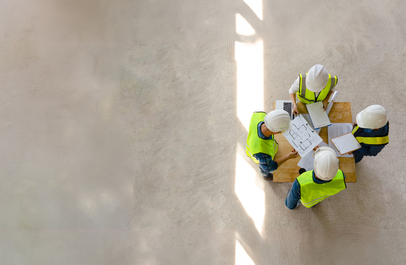 A project team wearing hard hats and high-visibility vests reviews building plans on site, supporting coordination for a commercial lighting or construction project.