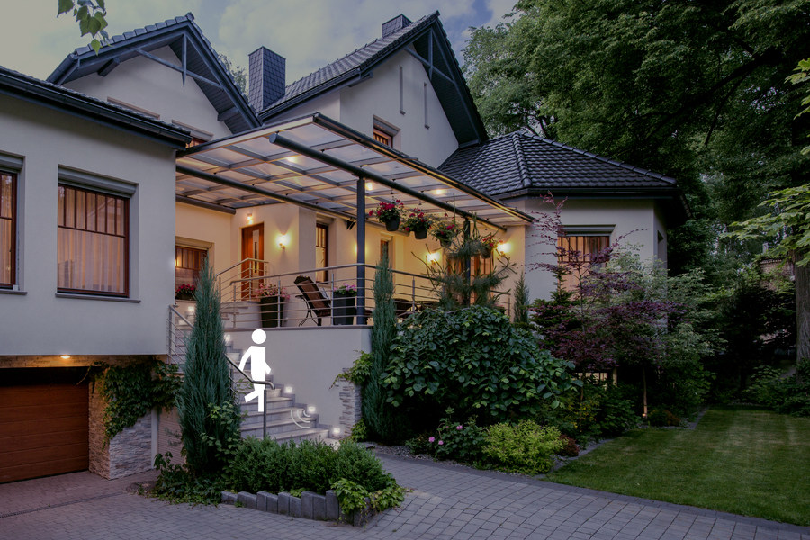 Extérieur résidentiel moderne au crépuscule avec un éclairage extérieur chaleureux illuminant l’entrée principale, le patio couvert, l’allée, l’aménagement paysager, les escaliers et la zone du garage.